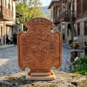 A high-definition photograph of the unique KÜP Ceramics House handcrafted 1-of-1 ceramic plate, showcasing a detailed 3D relief panorama of old Sheki streets and the historic mosque minaret, resting flat on a natural wooden display stand against a blurred background, certified authentic by master Kamran Ibrahimkhalilov.