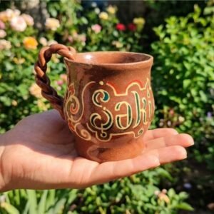 A high-definition front view photograph of the unique KÜP Ceramics House handcrafted 1-of-1 ceramic mug featuring intricate geometric Sheki carpet motifs and stylized Şəki calligraphy on a matte raw earthy clay body, with a detailed knitted-look tactile textured handle, certified authentic by master Kamran Ibrahimkhalilov.